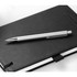 A silver ballpoint pen sits on top of a black notebook, featuring a smooth, click-action mechanism.