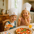 A kids chef hat in white, with a logo, worn by two children at a table with pizza and a drink.