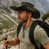 A wide-brimmed sun hat in dark green, worn by a man with a backpack, set against a mountainous landscape.