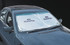 A silver sunshade with circular cutouts is placed on a car's windscreen, featuring a logo.