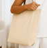 A neutral-toned tote bag held by a person, featuring a simple design and an attached logo.