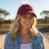A maroon cotton baseball cap with a curved peak, featuring a logo and worn by a woman with wavy blonde hair.