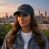 A vintage dark green baseball cap with a distressed look, featuring a logo and set against a city skyline at sunset.