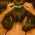 A drink cover scrunchie in vibrant green is being placed in a person's hair. The background features a blurred bar setting.