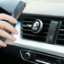 A clip-on car phone holder in silver attached to a car air vent, with a smartphone being positioned near it.