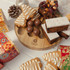 A bamboo platter with decorated gingerbread cookies, chocolate balls, and festive slices, surrounded by wrapping paper and ornaments.
