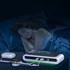 A wireless charging station in white and green, charging devices beside a sleeping person in a dimly lit bedroom.