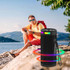 A black wireless speaker with colourful LED lights, positioned by a lake with a person in the background.