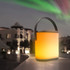 A portable LED speaker with a warm orange glow, featuring a handle, set against a scenic backdrop.
