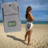 A grey eco-friendly bag displayed on a beach, featuring a logo, with a person wearing it while facing the ocean.