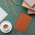 A5 PU leather notebook in brown with a logo, positioned beside a cup of coffee and various papers on a green background.