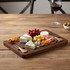 A slate cheeseboard with a wooden frame, featuring assorted cheeses, nuts, grapes, and garnishes. A glass of wine is nearby.