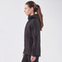 A women's softshell jacket in black, featuring a hood and zippered pockets, displayed on a model from the side.