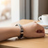 A sleek metal buckle latch bracelet in silver and black on a wrist, resting on a wooden table near a cup of coffee.