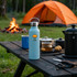 A drink bottle in light blue stainless steel with a logo, placed on a table near a tent and campfire.