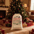 A large cream drawstring bag with a festive design, placed near a decorated Christmas tree and wrapped presents.