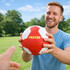 A red and white machine-stitched PU handball held by a smiling man in a grassy outdoor setting.
