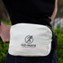 A beige cotton belt bag with a logo, worn on the waist against a green background.