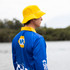 A man wearing a blue long sleeve fishing polo and a yellow bucket hat stands by the water. The shirt features a logo.