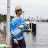 A man wearing a blue long sleeve polyester fishing tee stands by the water, surrounded by a city skyline and fishing gear.