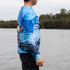 A long sleeve sublimated fishing tee in blue, featuring a fish design, displayed against a waterfront backdrop.