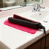A black hair straightener and a red styling tool rest on a pink silicone mat on a bathroom countertop.
