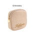 A beige mini makeup bag featuring a zip closure and gold embroidery, with a small round charm attached.