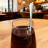 A stainless steel spoon hangs from a clear glass containing dark liquid, set on a wooden table in a café.