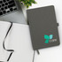 A grey notebook with a logo on the cover, lying open beside a laptop and a small plant.