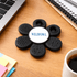 A black fidget spinner in a flower shape with a central logo, placed on a wooden desk near a coffee cup and stationery.