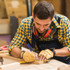 A man wearing a checkered shirt and gloves is measuring and marking wood in a workshop setting.