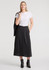 A woman stands wearing a white t-shirt and a black midi skirt, paired with ankle boots. The skirt features buttons and has a logo.