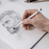 A hand holding a white pencil draws a sketch of a bearded man in a notebook. Pencils are visible in the background.