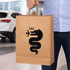 A brown paper show bag with a black logo featuring a snake. The bag is being held by a person in casual attire.