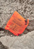An orange packable rain jacket neatly folded and resting on rocky terrain, featuring a logo and product details.