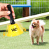 A yellow 75mm hand bell with a black handle, held above a small dog on grass. The bell has a logo.