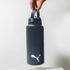 A drink bottle in dark blue with a handle and a logo on the front, suspended by a hand.