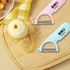 A wooden cutting board with a peeled pear and two peelers in pink and light blue, featuring a logo.