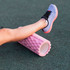 A pink foam yoga roller positioned on green flooring, with a person's leg resting on it, wearing blue running shoes.