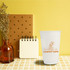 A white disposable double wall paper cup with a logo, beside a brown notebook and a vase of dried flowers.