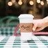 A white compostable paper cup with a brown cup sleeve held by a hand, set on a checkered table.