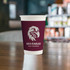 A single wall paper cup in maroon featuring a logo, set against a blurred background of a café environment.