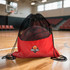 A red drawstring mesh backpack with a basketball inside, featuring a logo on the front, set on a wooden court.
