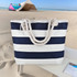 A striped tote bag in navy and white with a rope handle, set against a beach backdrop.
