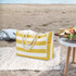 A yellow and white striped beach bag with rope handles, placed on a sandy surface near a picnic setup.