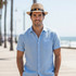 A man wearing a tan Madrid style string straw hat and a light blue shirt, with palm trees and the ocean in the background.