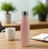 A drink bottle in pink with a stainless steel lid, set on a wooden table near a mug and a notebook.