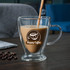 A transparent, V-shaped double wall mug filled with coffee, featuring a logo, sits on a wooden table with coffee beans nearby.