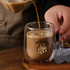 A barrel-shaped double wall mug in clear glass is being filled with a creamy coffee beverage.
