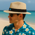 A sun hat in a light beige colour with a black band, worn by a man in a floral patterned shirt by the beach.